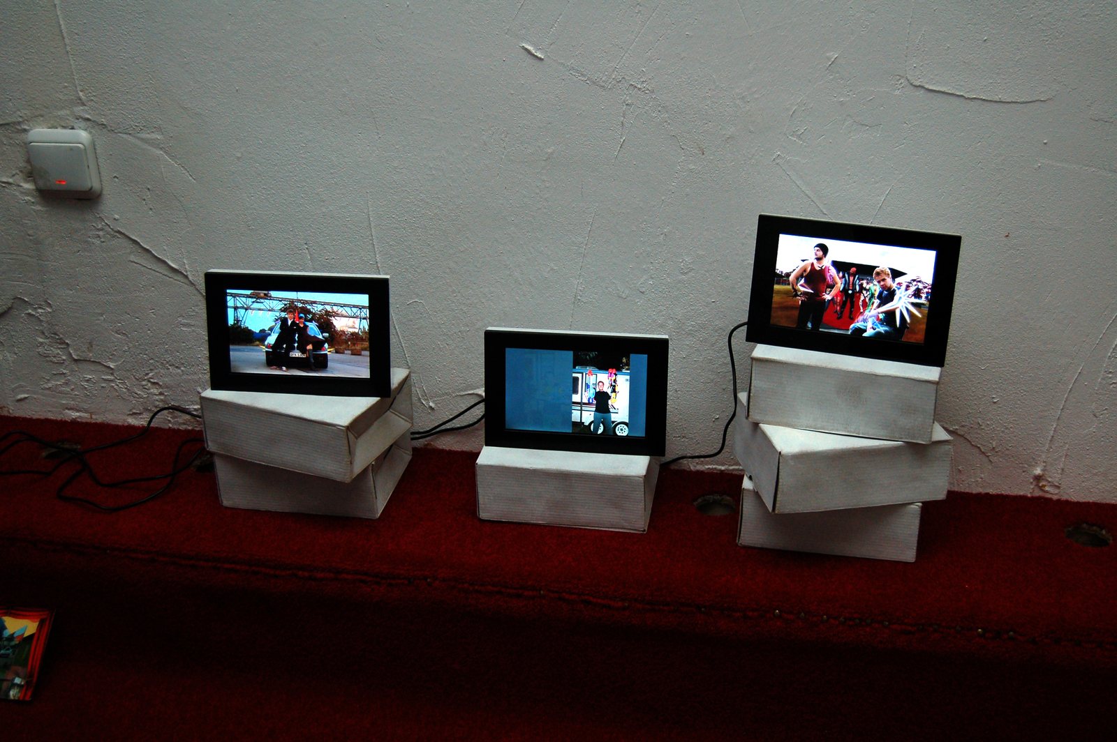 Three screens on white plinths on a red carpet, playing the hybridhappening recordings